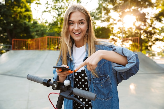 Foto di un'adolescente allegra positiva nel parco che cammina sullo scooter utilizzando il puntamento del telefono cellulare.