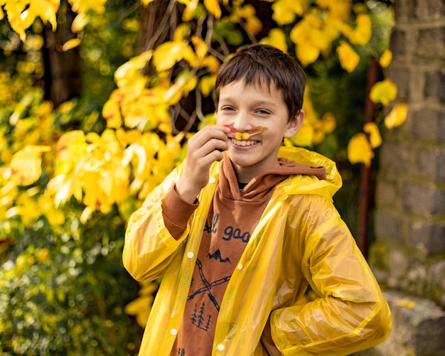 Foto di un adolescente con un impermeabile giallo tra le foglie autunnali, passeggiate nel parco, indulgenza, fa i baffi con foglie d'acero, foto umoristica