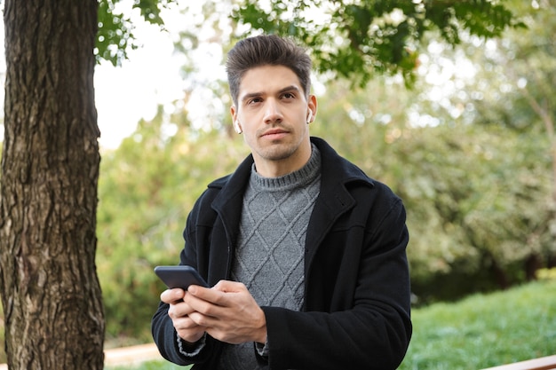 Foto di un bel giovane serio in abbigliamento casual che cammina all'aperto nel parco verde utilizzando la musica d'ascolto del telefono cellulare con gli auricolari.