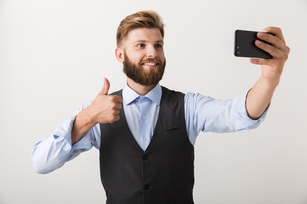 Foto di un bel giovane uomo barbuto in piedi isolato sopra il muro bianco prendere un selfie dal telefono cellulare.