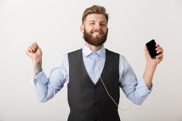 Foto di un bel giovane uomo barbuto in piedi isolato sopra la parete bianca che tiene musica d'ascolto del telefono cellulare.