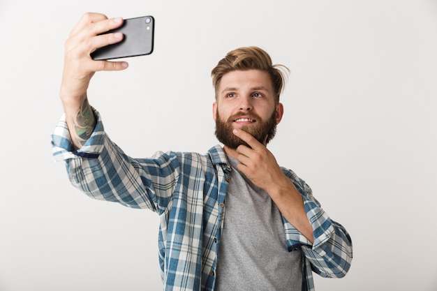 Foto di un bel giovane uomo barbuto in piedi isolato su sfondo bianco muro prendere un selfie dal telefono cellulare.