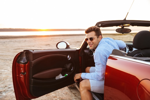 Foto di un bell'uomo caucasico che indossa occhiali da sole che esce da un'elegante auto convertibile in riva al mare all'alba