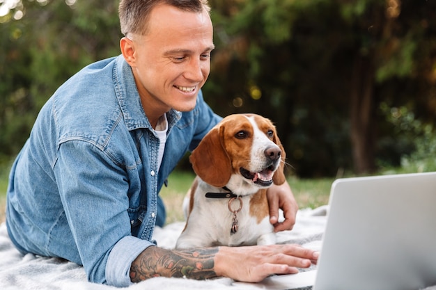 Foto di un giovane caucasico sdraiato su una coperta nel parco con il suo cane canino e il computer portatile