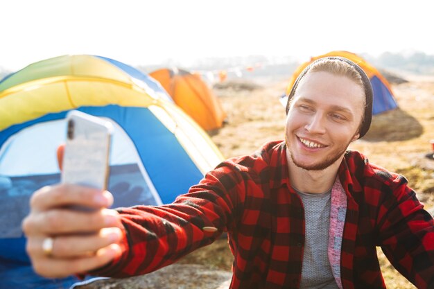 Foto di un giovane fuori in una vacanza alternativa gratuita in campeggio sulle montagne usando il telefono cellulare per fare un selfie.