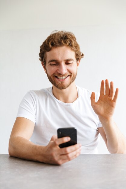 Foto di un giovane uomo barbuto felice al tavolo a casa parlando da telefono cellulare agitando.