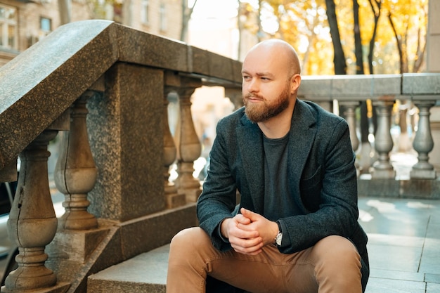 Foto di un giovane uomo barbuto seduto sulle scale nella vecchia città europea e distogliendo lo sguardo