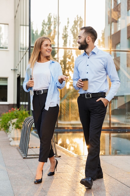 Foto di un giovane uomo d'affari allegro sorridente felice e donna d'affari fuori in strada vicino al centro affari che cammina tenendo appunti e tazza di caffè che parlano tra loro.