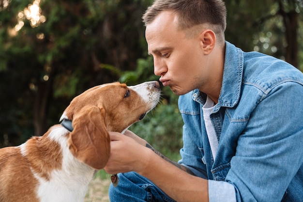 Foto di un giovane uomo felice in abiti di jeans che bacia il suo cane mentre cammina nel parco