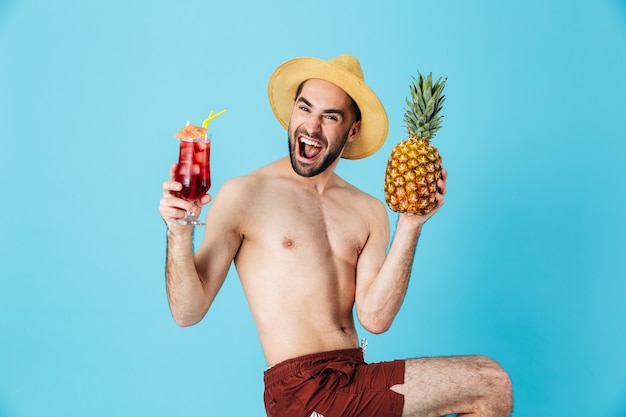 Foto di un turista caucasico senza camicia che indossa un cappello di paglia che sorride mentre tiene in mano ananas e cocktail isolati