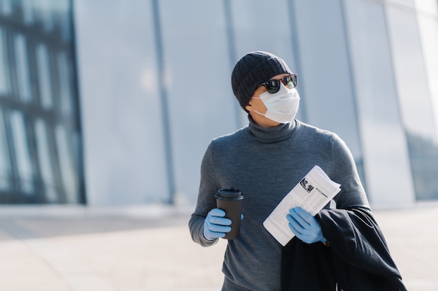 Foto di un uomo adulto che cammina in strada, indossa una maschera protettiva e guanti di gomma come protezione contro le malattie infettive, beve caffè da asporto, ha comprato il giornale per leggere le notizie, concentrato a parte