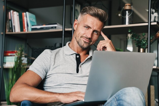 Foto di un uomo adulto felice in abbigliamento casual che usa il laptop mentre è seduto sulla poltrona in appartamento