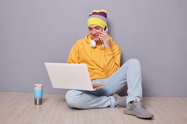 Foto di un uomo allegro sorridente che indossa un cappello a cuffia con cappuccio giallo e jeans che lavora su un notebook che parla su uno smartphone e sorride con una piacevole conversazione in posa contro il muro grigio