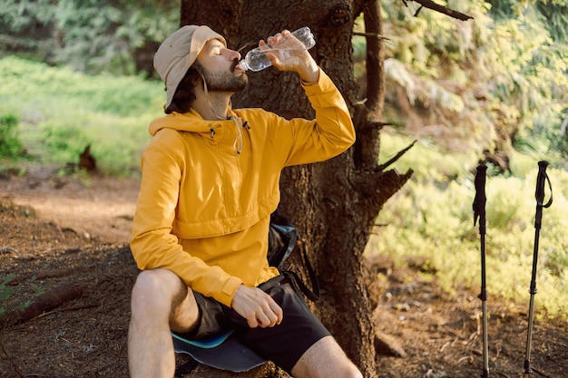 foto di un uomo assetato che viaggia tra le montagne boscose dei Carpazi e beve acqua fresca da una bottiglia all'ombra di un albero con grande sete