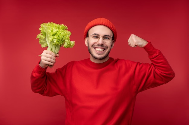 Foto di un uomo attraente con la barba in occhiali e vestiti rossi. Il maschio tiene insalata, mostra forte e ama le verdure, isolate su sfondo rosso.