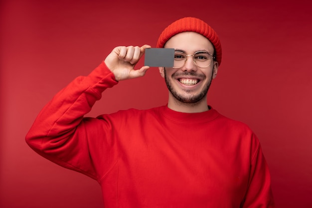 Foto di un uomo attraente con la barba in occhiali e vestiti rossi. L'uomo felice tiene la carta di credito davanti al viso, isolato su sfondo rosso.