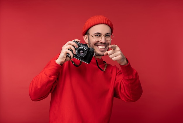 Foto di un uomo attraente con la barba in occhiali e vestiti rossi. L'uomo felice tiene la macchina fotografica e sembra bello, isolato su sfondo rosso.
