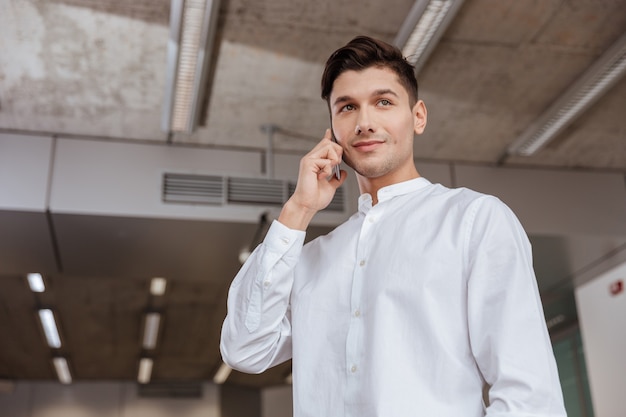 Foto di un uomo attraente vestito con una camicia bianca che parla al telefono al chiuso. Coworking. Guardando da parte.