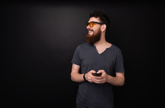 Foto di un uomo barbuto che tiene in mano un telefono cellulare mentre guarda lontano, su sfondo grigio scuro