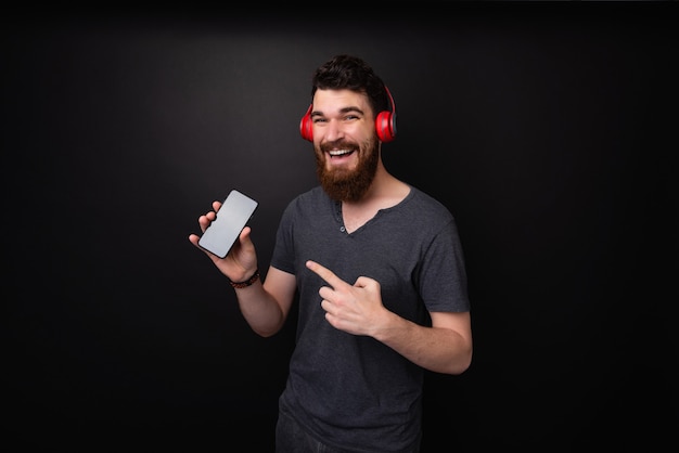 Foto di un uomo barbuto con le cuffie che puntano al cellulare mentre guarda la telecamera su uno sfondo scuro