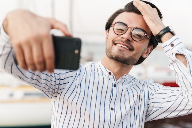 Foto di un uomo brunetta felice con gli occhiali che scatta una foto selfie sullo smartphone e sorride mentre cammina sul molo