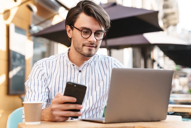 Foto di un uomo caucasico concentrato che indossa occhiali usando laptop e cellulare mentre lavora al bar all'aperto