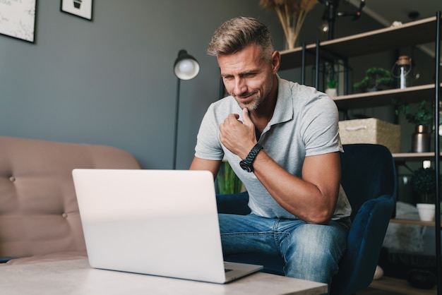 Foto di un uomo caucasico di successo in abbigliamento casual che usa il laptop mentre è seduto sulla poltrona di casa