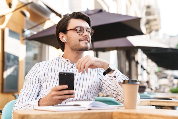 Foto di un uomo caucasico sorridente che indossa occhiali usando auricolari e cellulare mentre lavora al bar all'aperto