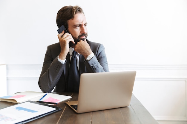 Foto di un uomo d'affari barbuto bello e serio che si siede in ufficio utilizzando un computer portatile che lavora al chiuso parlando al telefono cellulare.