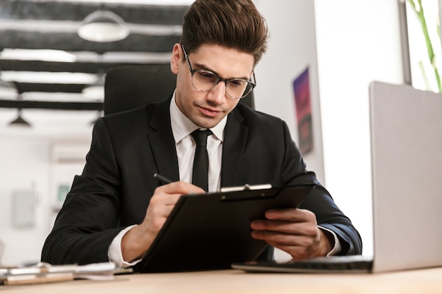 Foto di un uomo d'affari serio con gli occhiali che lavora con documenti e laptop mentre è seduto alla scrivania in un ufficio a pianta aperta