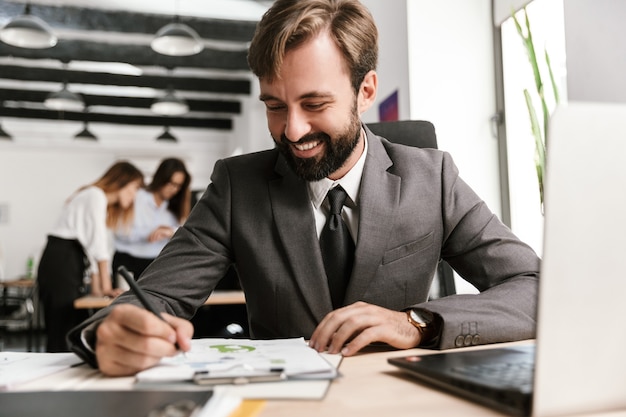 Foto di un uomo d'affari sorridente in abito formale che lavora con documenti e laptop mentre è seduto alla scrivania in un ufficio a pianta aperta