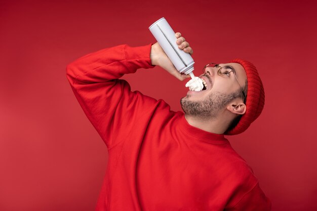 Foto di un uomo felice con la barba in occhiali e vestiti rossi. Divertiti a mangiare la panna, isolata su sfondo rosso.