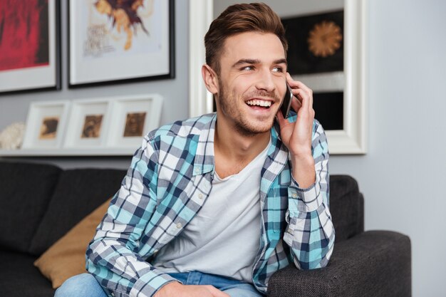 Foto di un uomo gioioso vestito con una camicia in una stampa a gabbia seduto sul divano di casa e parlando con il suo telefono.