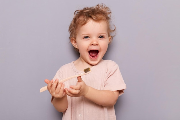 Foto di una bambina carina che tiene lo spazzolino da denti provando a lavarsi i denti al mattino ogni giorno in piedi isolato su sfondo grigio Assistenza medica sanitaria