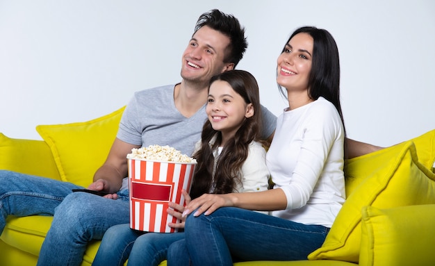 Foto di una bambina carina e dei suoi genitori, che mangiano popcorn insieme, mentre guardano programmi TV sul divano.