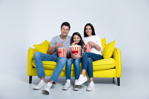 Foto di una bambina carina e dei suoi genitori, che mangiano popcorn insieme, mentre guardano programmi TV sul divano.