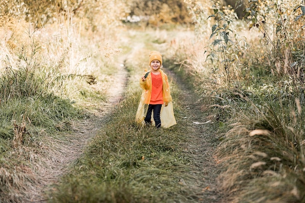 Foto di una bambina divertente con cappello giallo e impermeabile che cammina lungo la strada tra l'erba alta e sorride