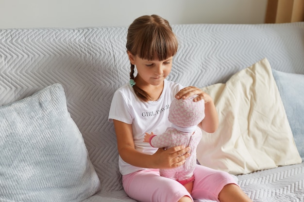 Foto di una bambina in età prescolare seduta sul divano nella stanza di casa che gioca con una bambola come mamma, bambino che indossa una maglietta bianca e pantaloncini rosa, infanzia.