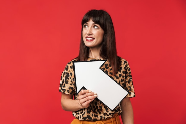 Foto di una bella donna bruna anni '30 vestita con abiti eleganti che sorride e tiene in mano un puntatore a freccia bianco isolato su un muro rosso