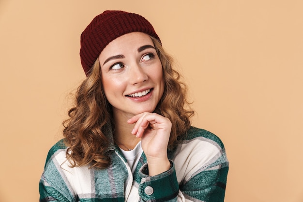 Foto di una bella donna contenta con un cappello a maglia che sorride e guarda da parte isolata sul beige