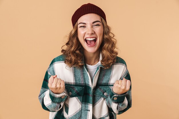 Foto di una bella donna gioiosa con un cappello a maglia che ride e fa il gesto del vincitore isolato sul beige