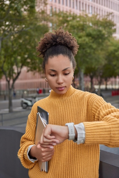 Foto di una bella giovane donna che indossa un maglione lavorato a maglia giallo controlla il tempo sull'orologio da polso tiene tablet e quaderni attende fino all'inizio delle lezioni pone all'aperto