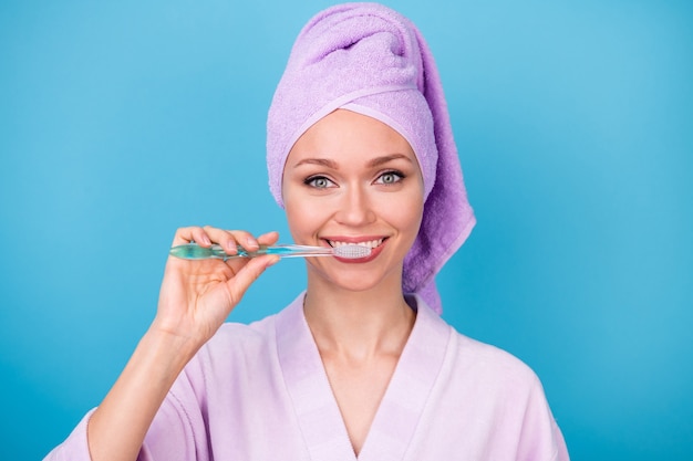 Foto di una bella giovane donna che si lava i denti mentre si prepara a indossare un asciugamano viola con turbante, accappatoio, sfondo di colore blu isolato