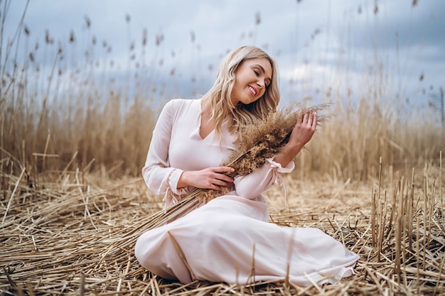 Foto di una bella ragazza sorridente con lunghi capelli biondi ricci in luce lunghi drees seduto in un campo di canna