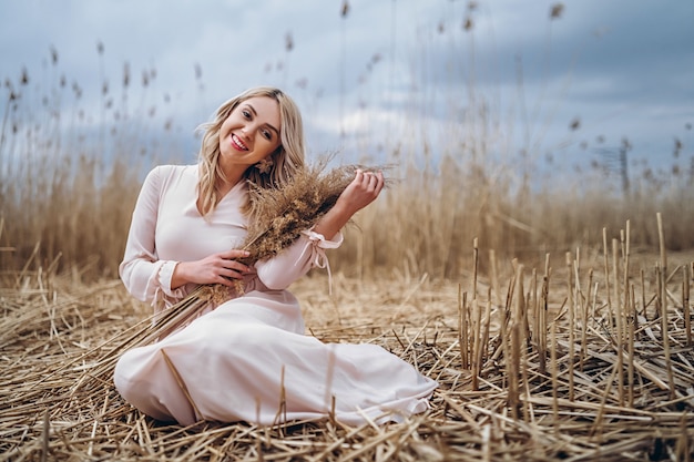 Foto di una bella ragazza sorridente con lunghi capelli biondi ricci in luce lunghi drees seduto in un campo di canna