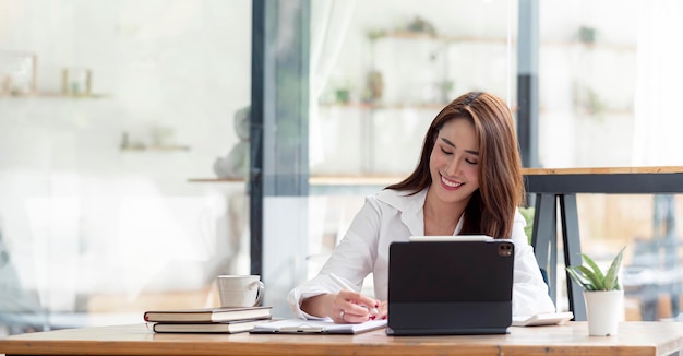 Foto di una donna abbastanza asiatica allegra che è stata impiegata per lavorare come dirigente sorridente seduto al desktop con un laptop e scrivendo una nota.