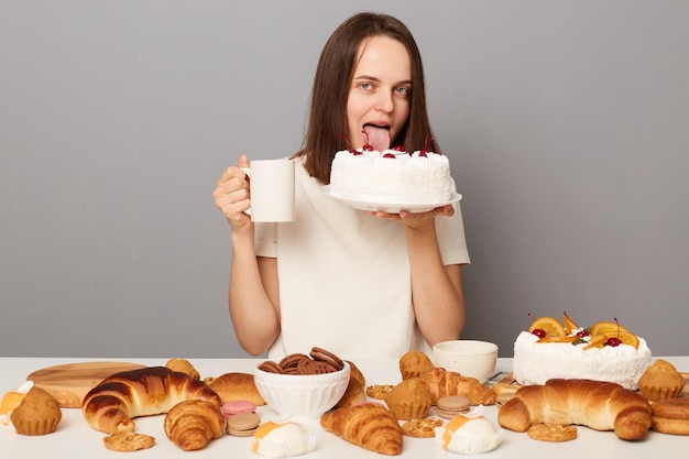Foto di una donna affamata con i capelli castani che indossa una maglietta bianca seduta al tavolo isolata su uno sfondo grigio che beve tè e lecca la torta guardando la fotocamera