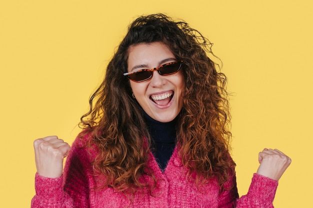 Foto di una donna allegra con i capelli ricci che stringe i pugni celebrando il successo indossando occhiali da sole isolati su sfondo giallo Persone e felicità