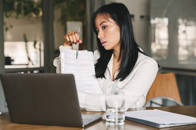 Foto di una donna asiatica di 20 anni che indossa abiti da ufficio tenendo e mostrando documenti cartacei sullo schermo del laptop, durante la videochiamata o la conferenza online