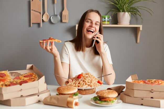 Foto di una donna attraente allegra e gioiosa con i capelli castani che indossa una maglietta bianca seduta in cucina a tavola e parla allo smartphone ridendo allegramente cenando mangiando pizza